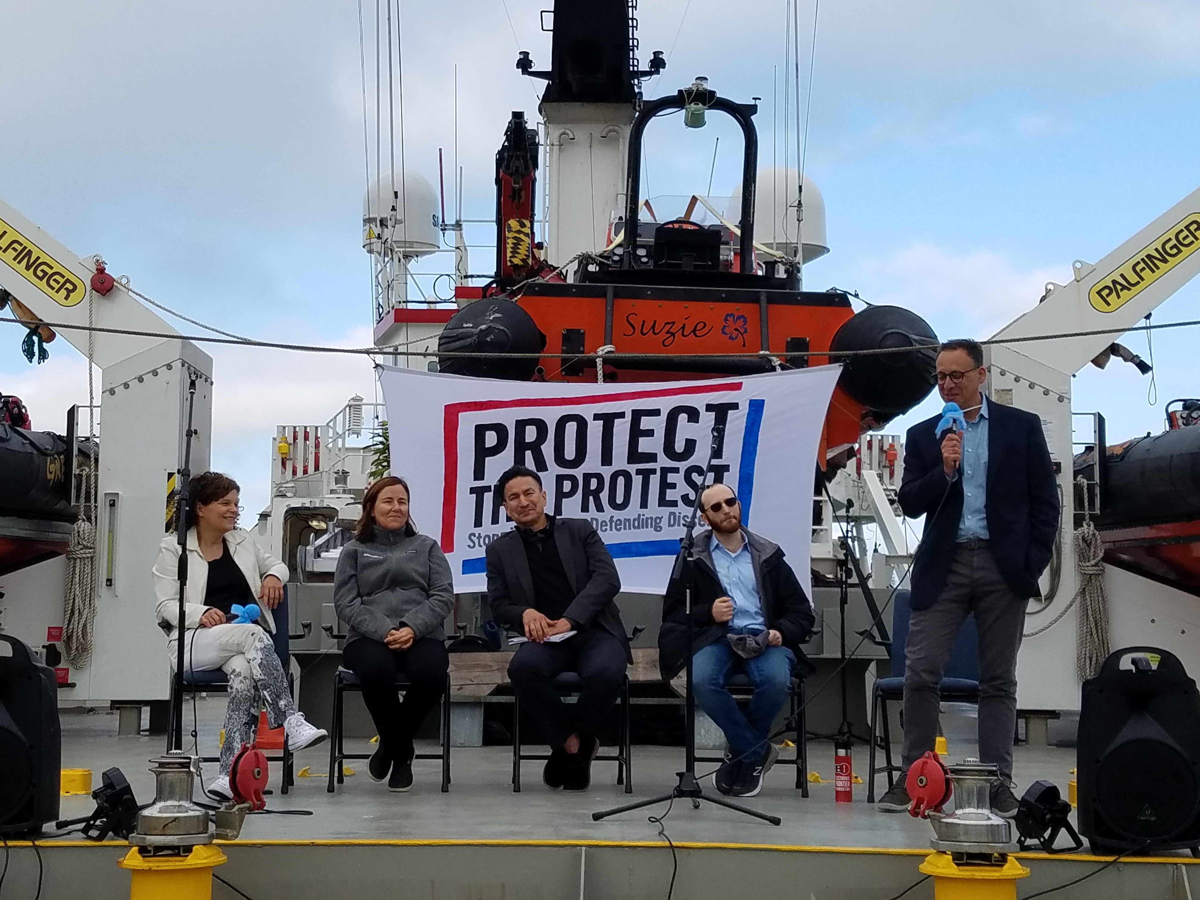 Aboard a ship that is carrying three smaller boats, four people face an audience to discuss their civil liberties work. A banner that says "Protect the Protest" hangs behind the speakers.