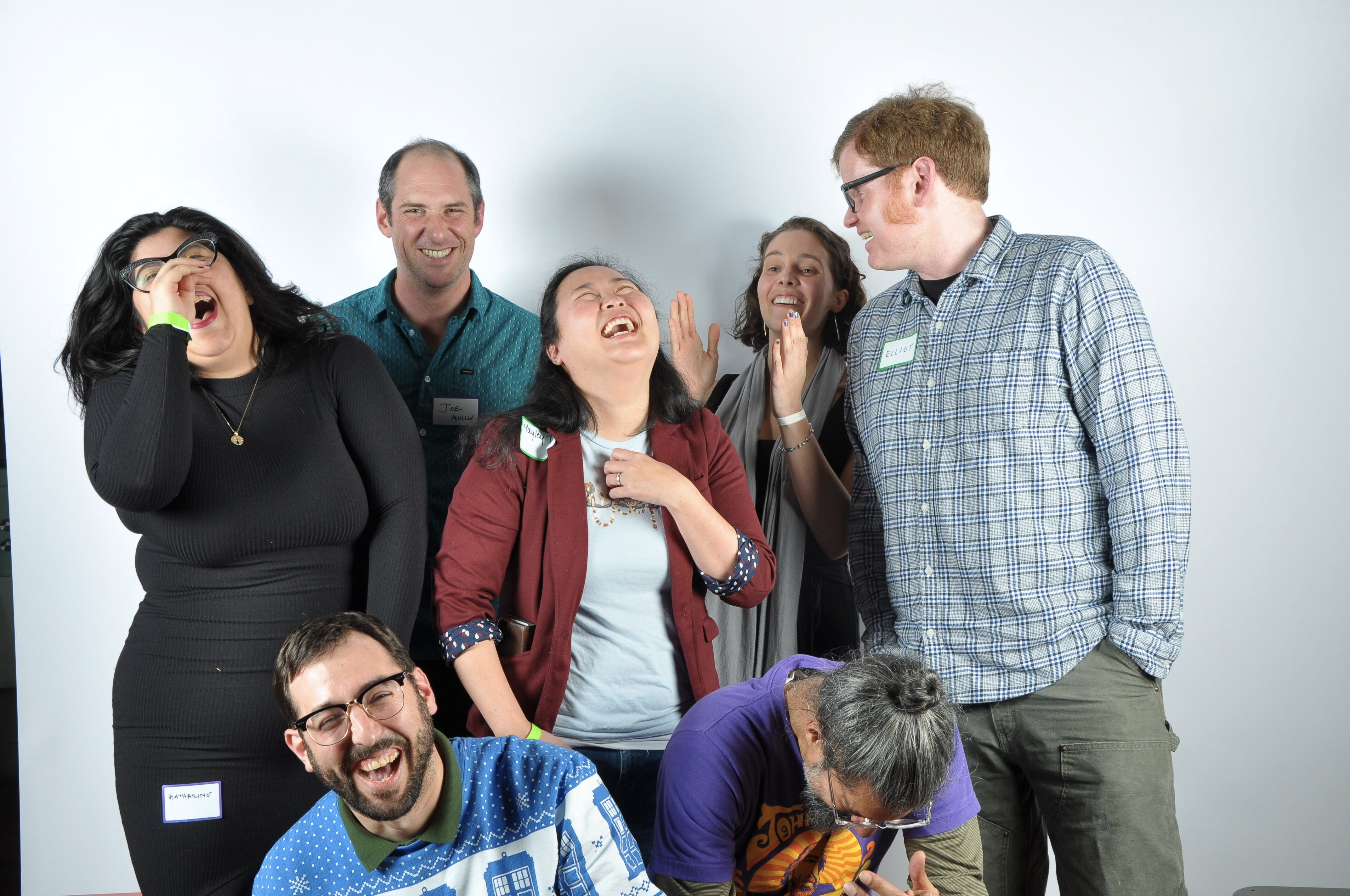 Elliot is turned to the side, speaking to the other six people, who are laughing. He has red hair, and is wearing glasses and a flannel shirt.