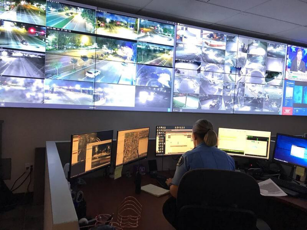 A police analyst sits at a workstation in front of a wall of video monitors. 