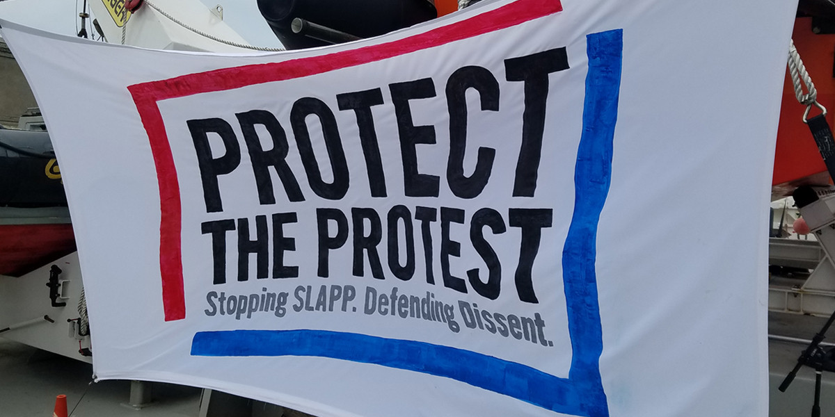 A white banner with black text that reads "Protect the Protest. Stopping SLAPP. Defending Dissent." The words are framed by a blue and red border on the banner. 